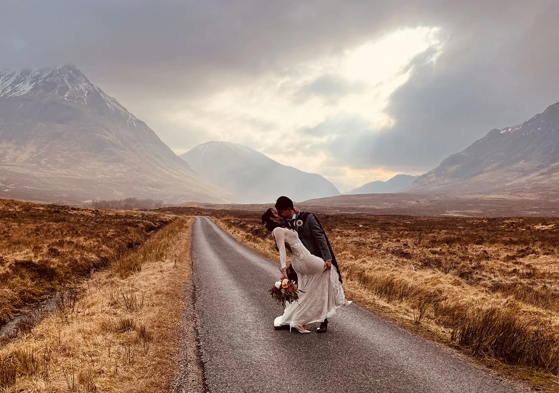 Groom dips bride holding bouquet in kiss with snow covered mountain tops in background